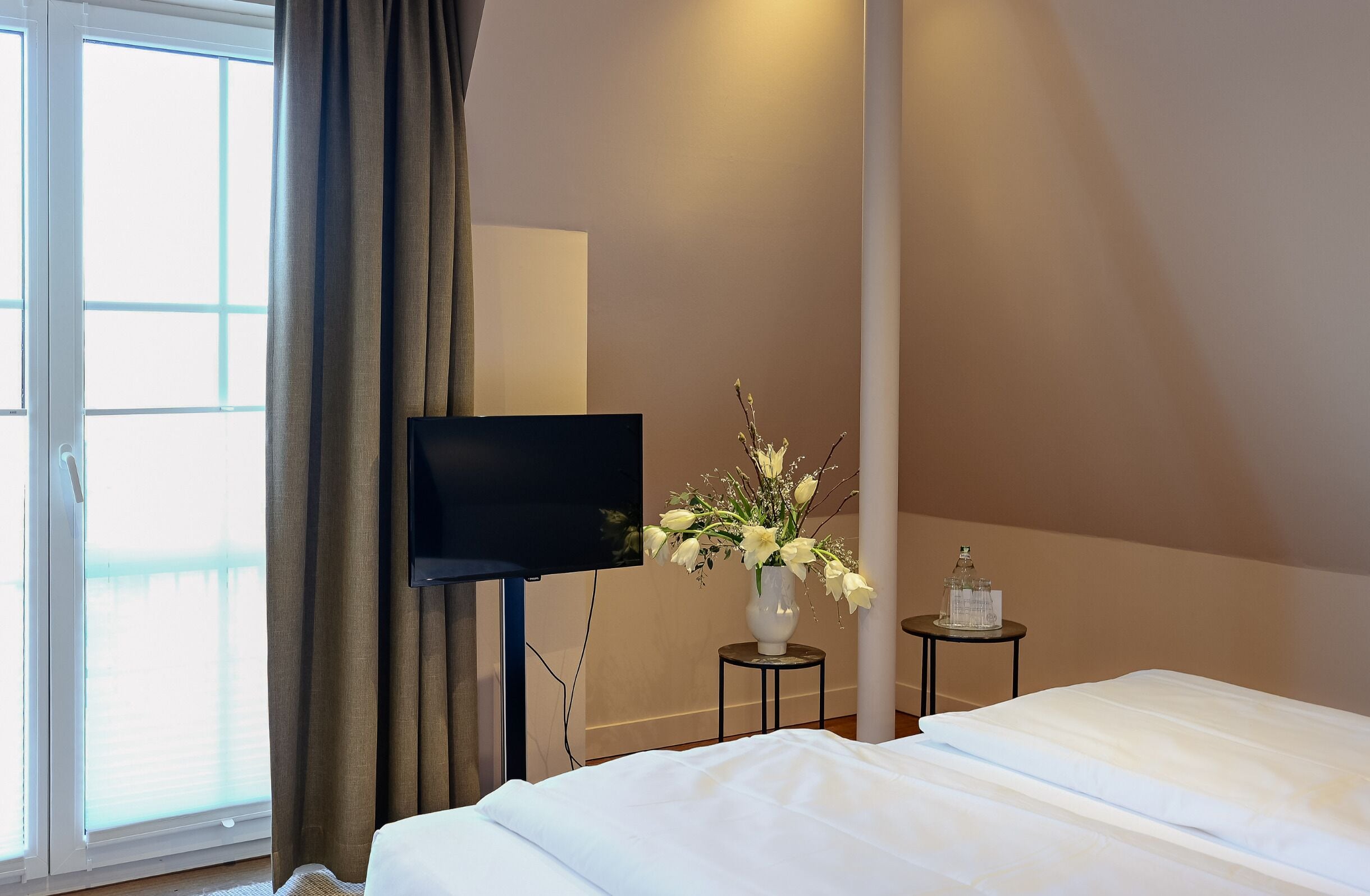 A hotel room with a bed, a TV, a vase with flowers, and a small table with a bottle.