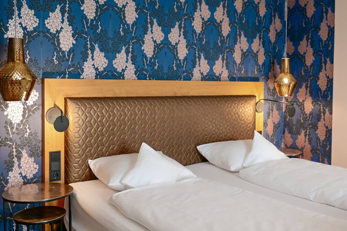 Una habitación de hotel con una cama, dos lámparas de noche, una mesa redonda y un papel tapiz estampado.