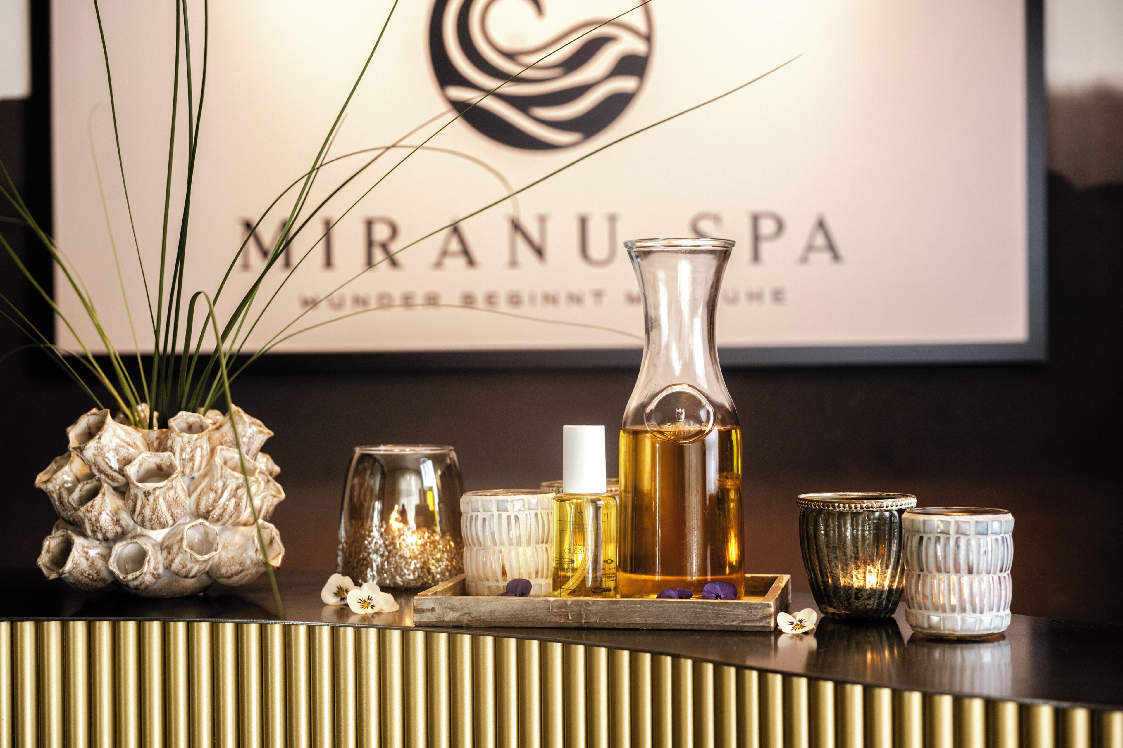 A spa setting with a framed sign, a vase with flowers, a bottle, and candles on a wooden tray.