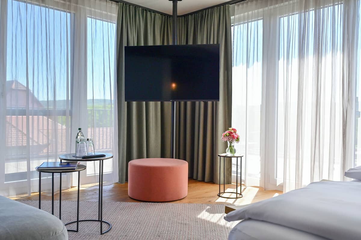 Una habitación de hotel moderna con un televisor de pantalla plana grande, un otoman redonda, una mesita redonda y un florero con flores.