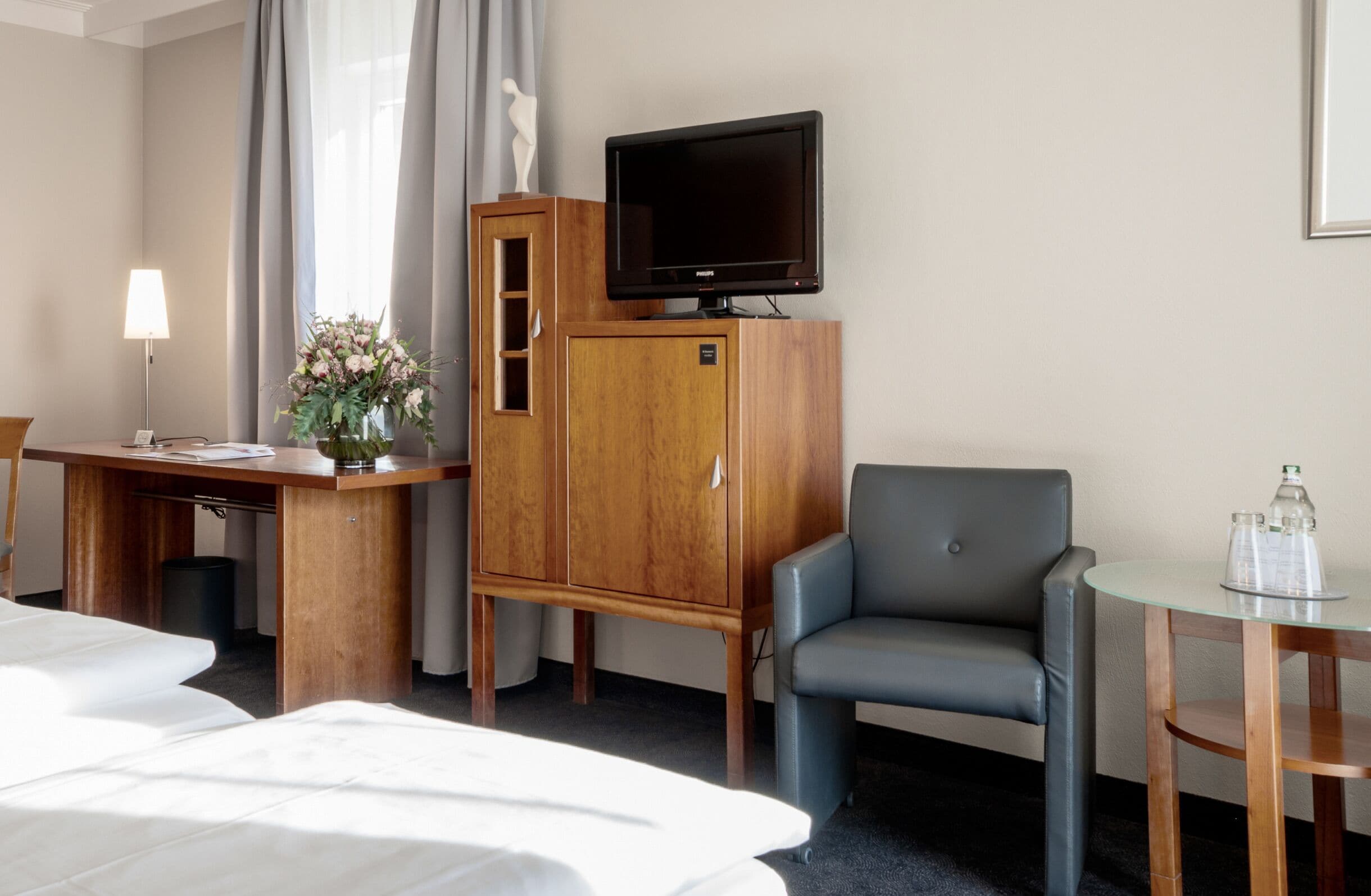 Une chambre d’hôtel avec un lit, un bureau doté d’une lampe, un téléviseur posé sur une commode en bois, un fauteuil gris et une petite table sur laquelle se trouve une carafe en verre.