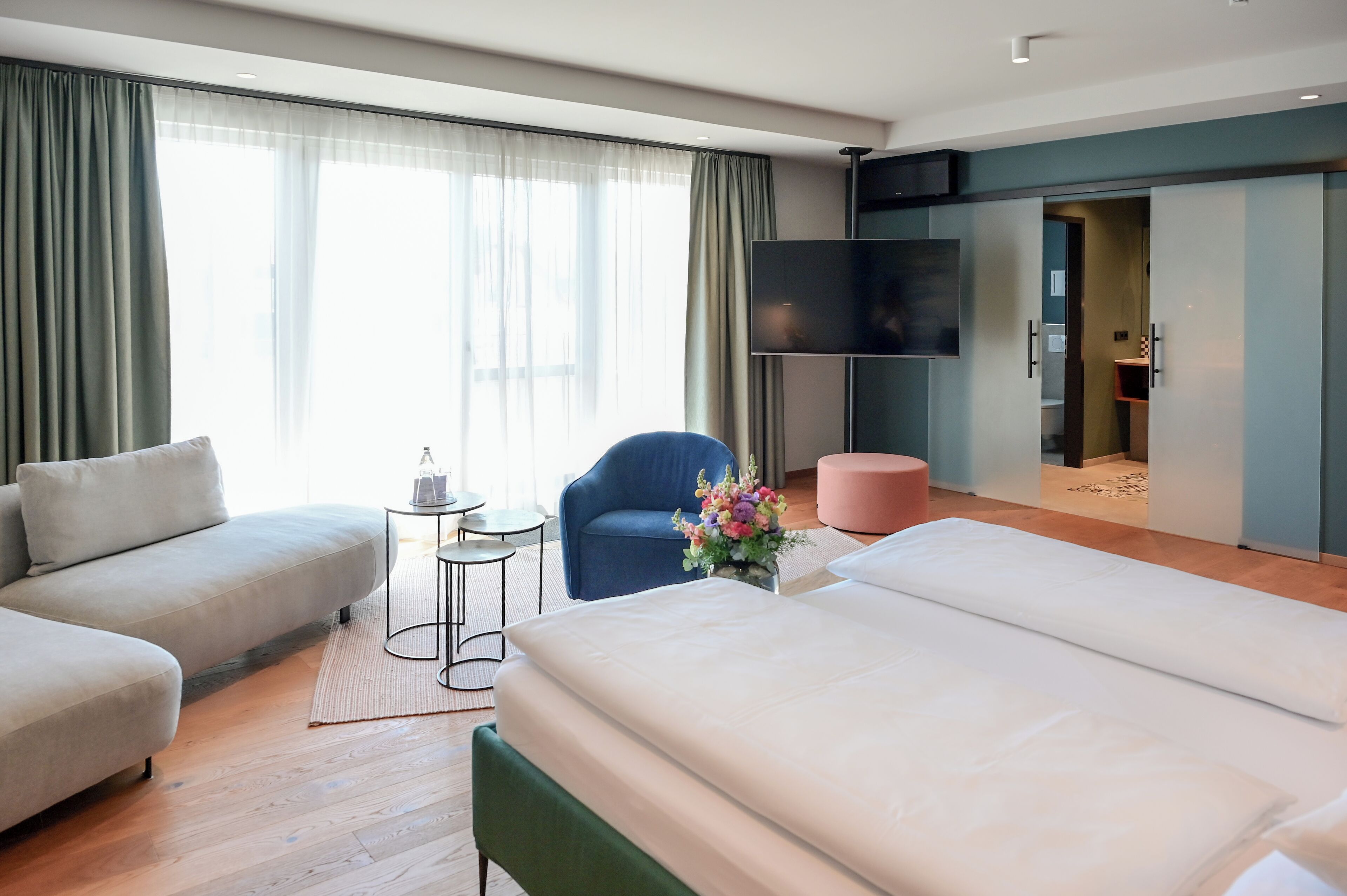 A modern hotel room with a sofa, a chair, a bed, a TV, and a small table with a vase of flowers.
