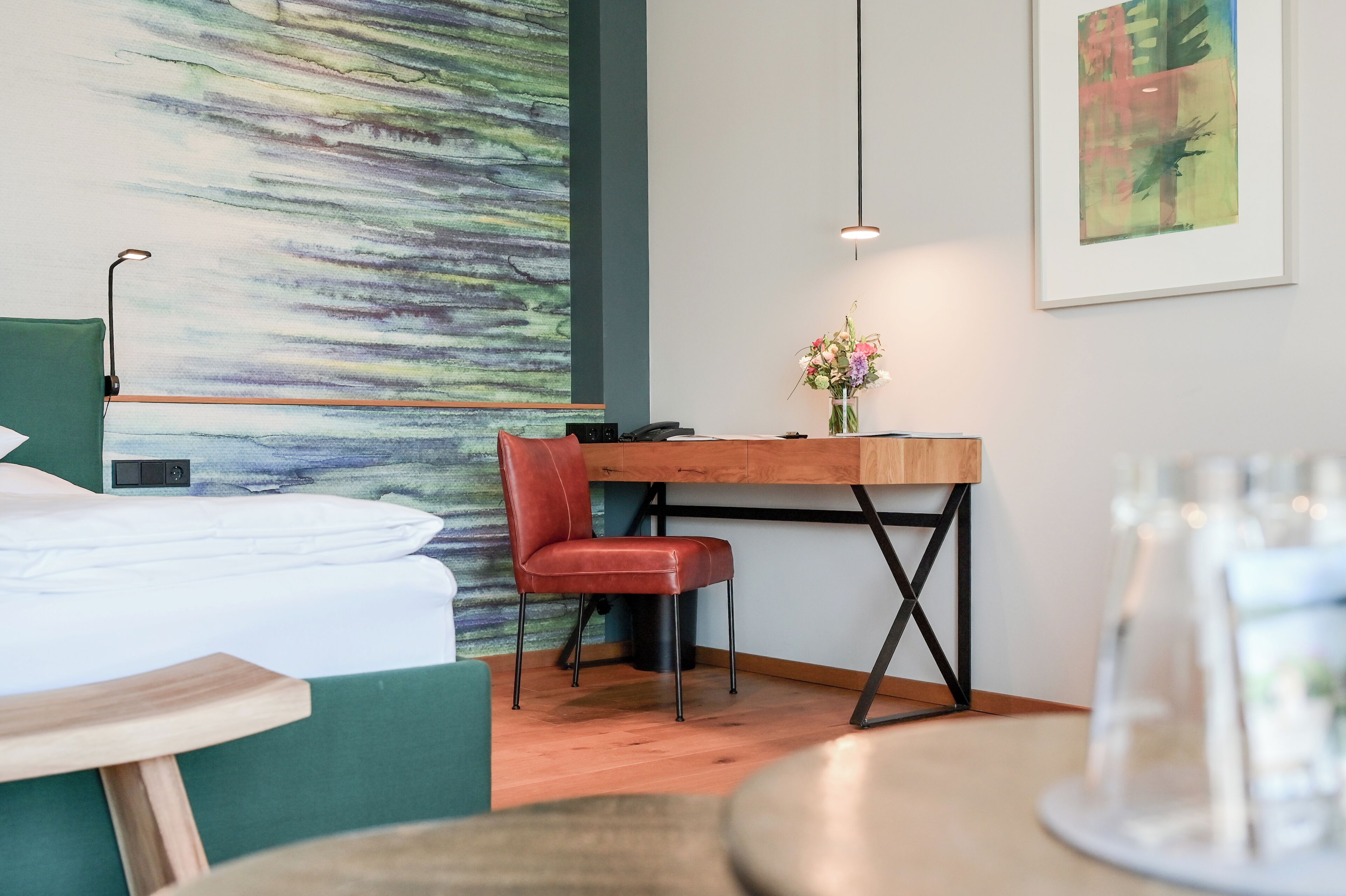 A hotel room with a bed, a desk with a chair, a vase with flowers, a painting on the wall, and a hanging light fixture.