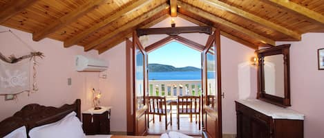Un dormitorio con techo de madera, una cama con ropa de cama blanca, un aparador de madera y vista al mar a través de una puerta de balcón.