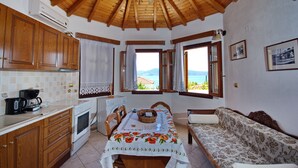 A kitchen with wooden cabinets, a dining table with a floral tablecloth, and a view of the sea through open windows. - Pelagos Villas (Alonissos)