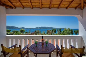 Una mesa de madera con dos vasos verdes y una botella, sobre un balcón con barandilla blanca, con vistas a un paisaje costero con montañas y el mar.