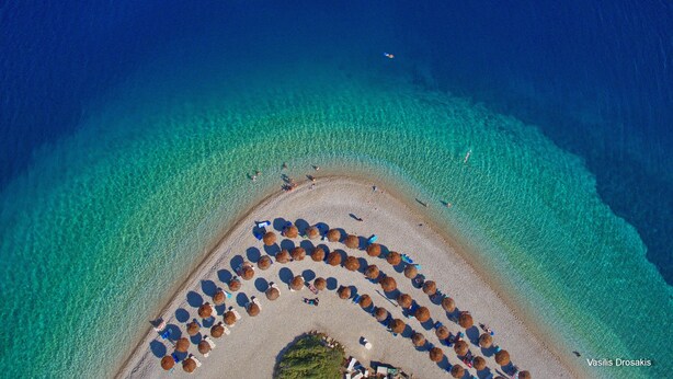 An aerial view of a beach with a circular arrangement of umbrellas and beach chairs, clear turquoise water, and a few people visible. - Pelagos Villas (Alonissos)