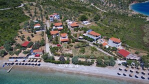 An aerial view of a coastal resort with multiple buildings, a beach with sun loungers, and clear turquoise waters. - Pelagos Villas (Alonissos)