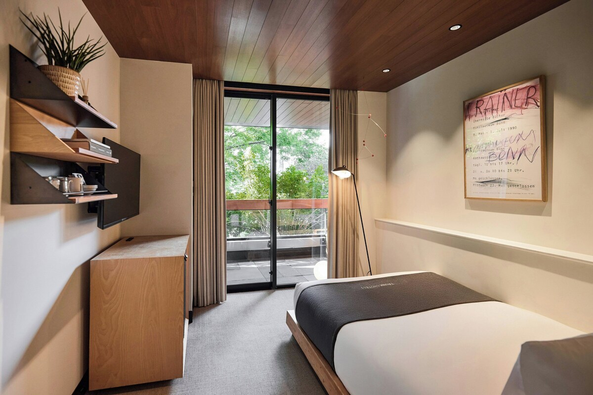 A modern hotel room with a bed, a wooden desk, a shelf with a plant, and a balcony with curtains.