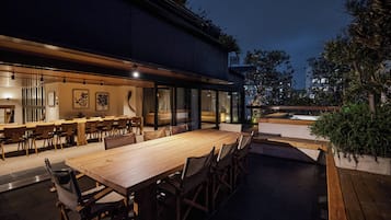 Un área de comedor al aire libre con una larga mesa de madera y sillas, rodeada de paredes de vidrio y plantas en macetas.