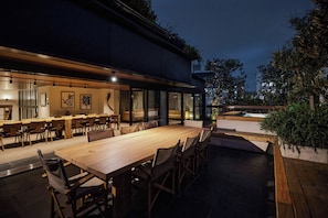 Un área de comedor al aire libre con una larga mesa de madera y sillas, rodeada por paredes de vidrio.