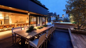 Un área de comedor al aire libre con mesas y sillas de madera, rodeada de plantas y vistas a la ciudad.