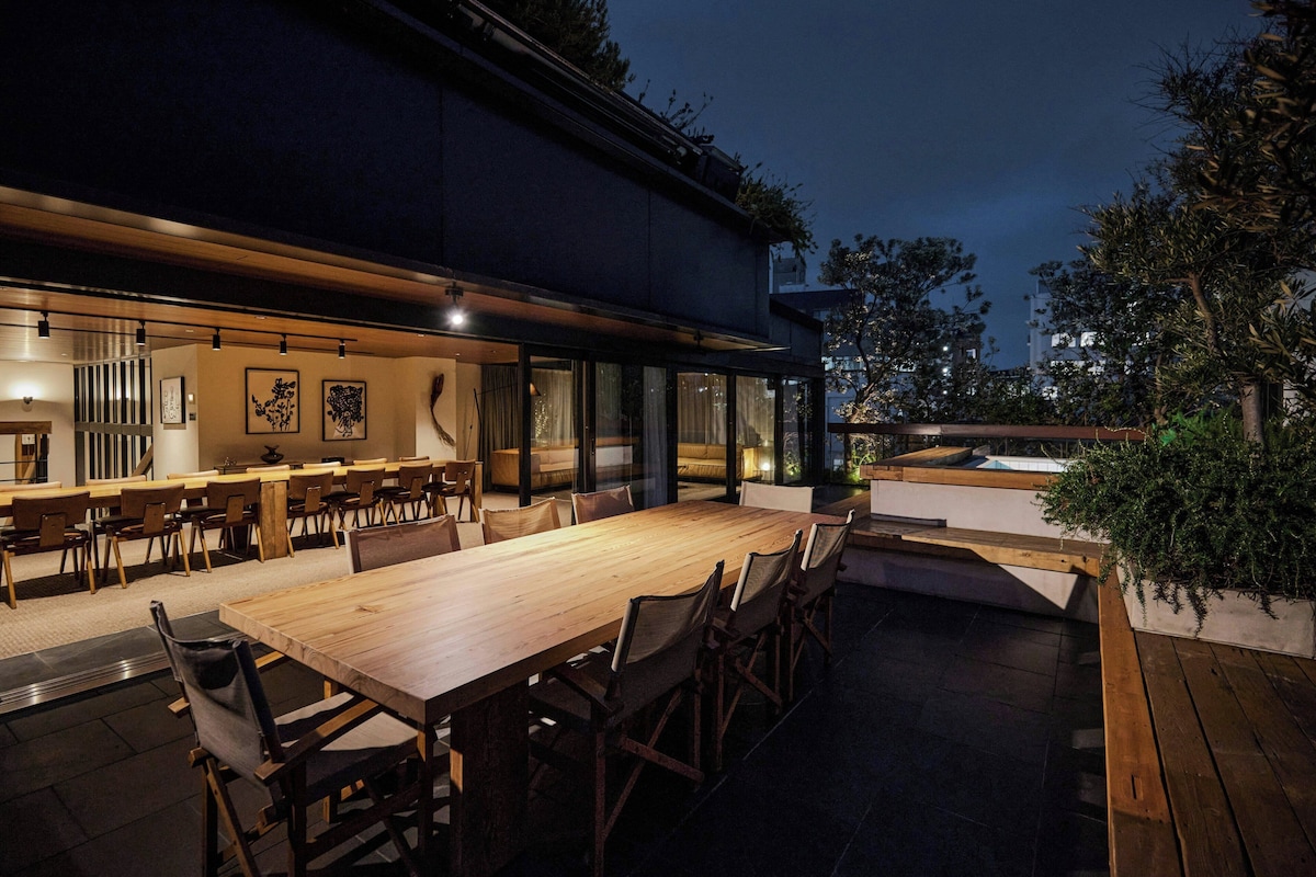 An outdoor dining area with a long wooden table and chairs, surrounded by a low wall with windows.