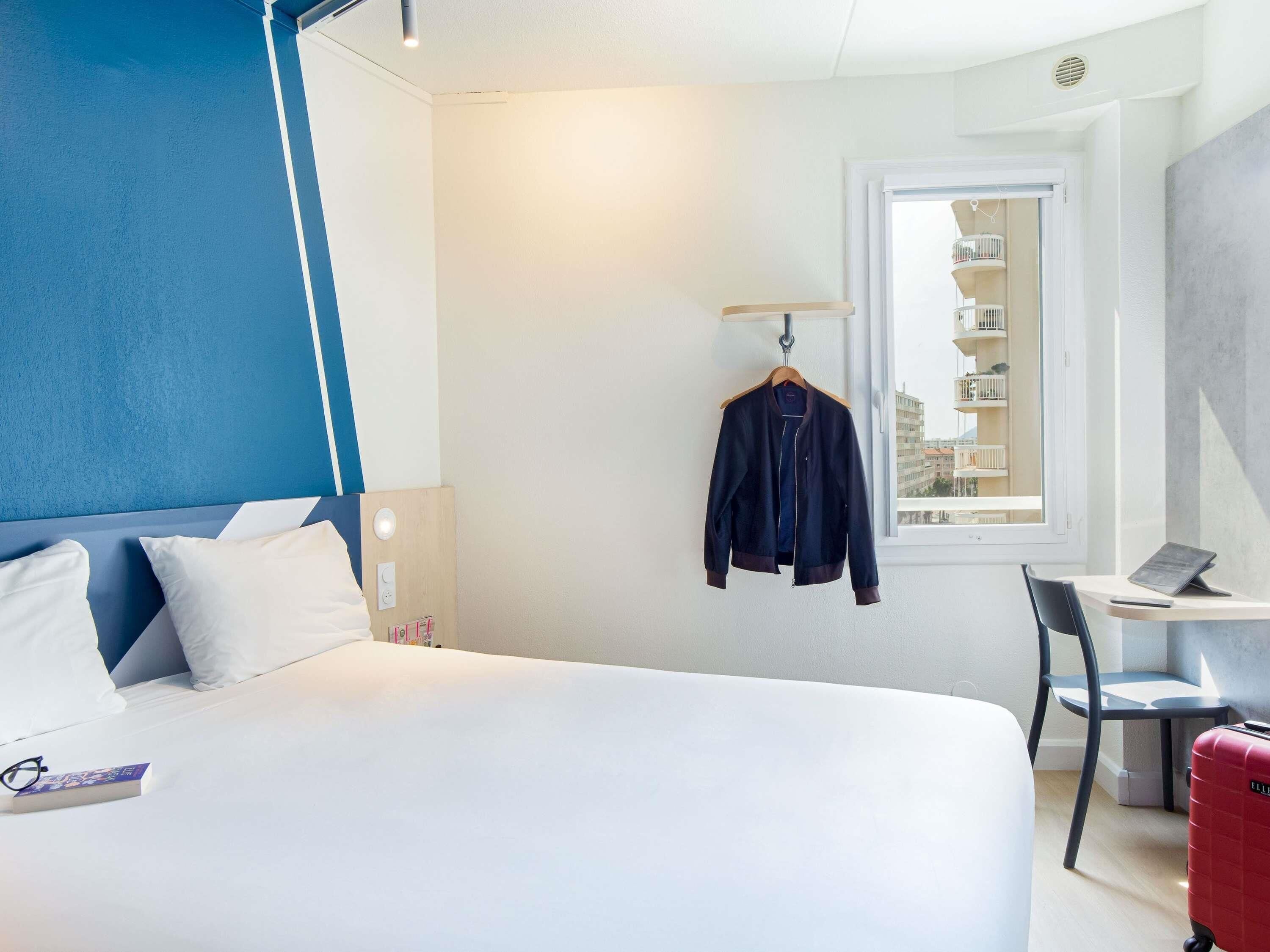 A hotel room with a bed, a desk, a chair, a hanging jacket, and a suitcase.