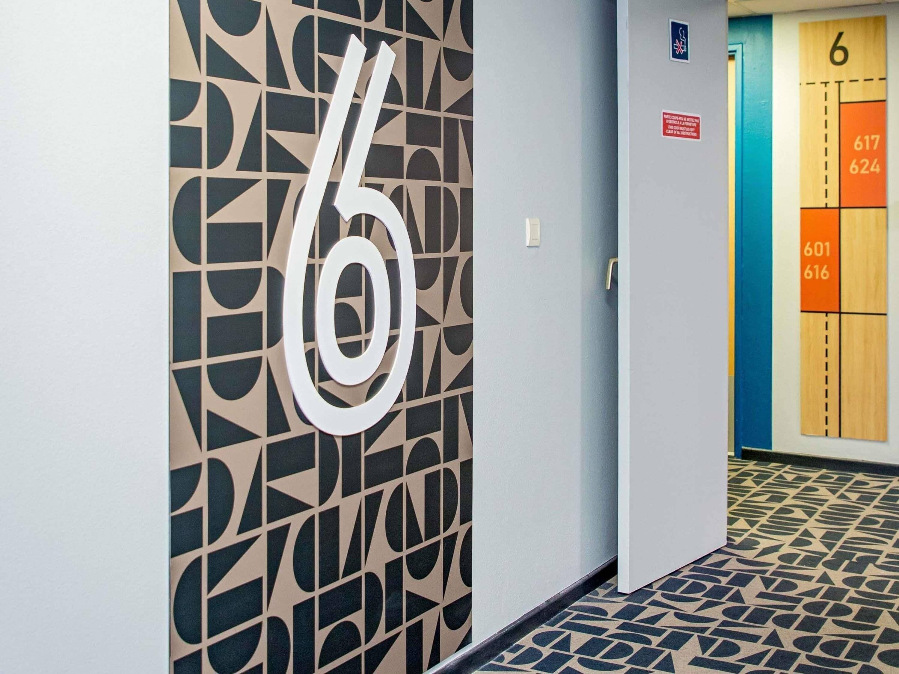 Un pasillo con un gran número 6 en la pared, una puerta con el número 6 y un suelo de estampado geométrico.