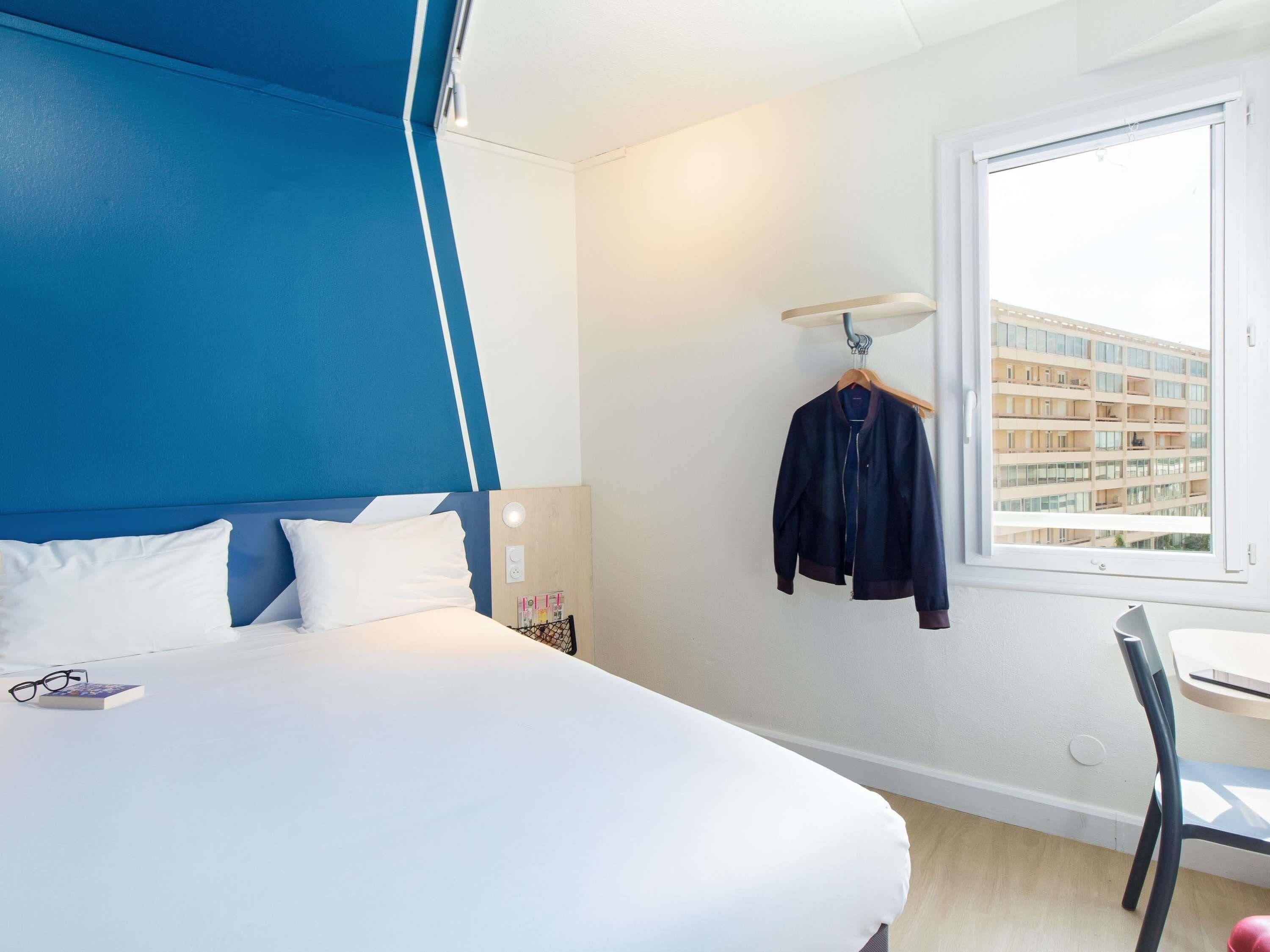 Une chambre d’hôtel avec un lit, un bureau, une chaise, une fenêtre donnant sur des immeubles et une veste accrochée.