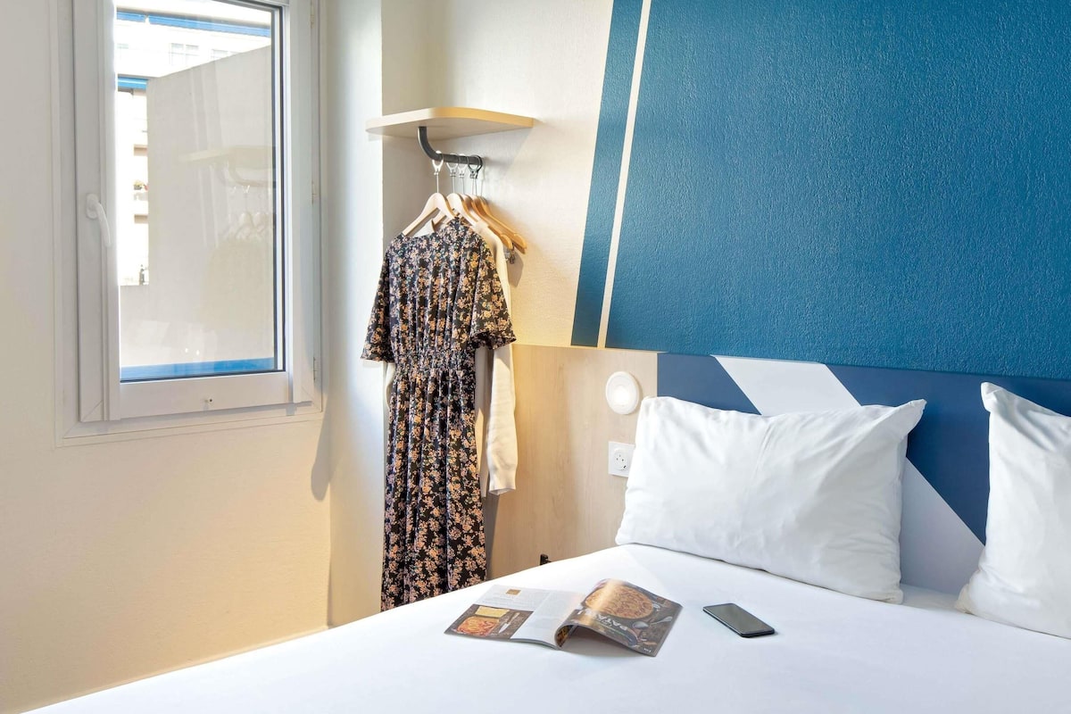 Hotelkamer met een bed, een raam, een wandlamp, een hangende kledingstuk en een tijdschrift op het bed.