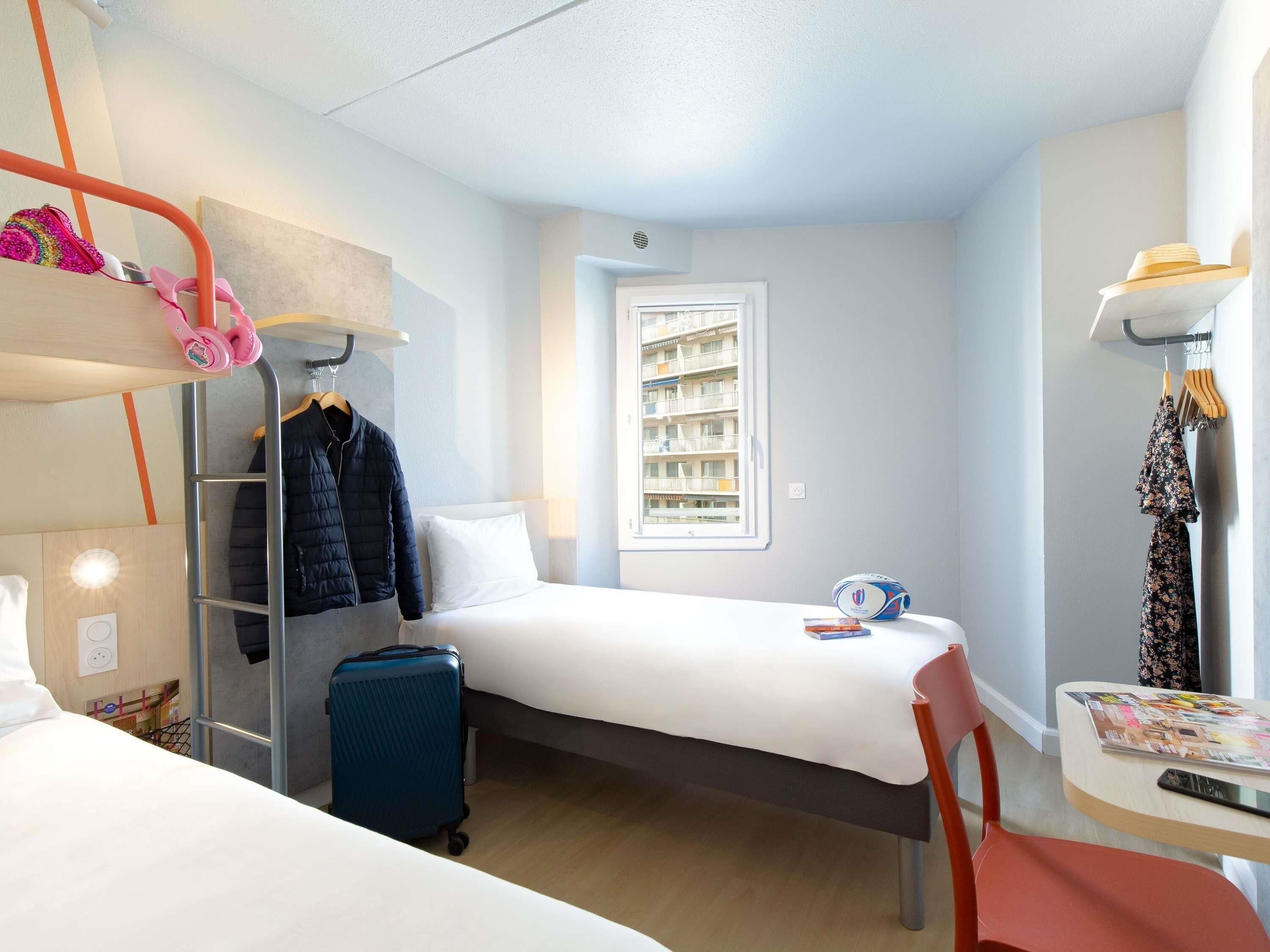 Ett litet hotellrum med en våningssäng, en enkelsäng, en resväska, ett bord med ett tidningsnummer och ett fönster med utsikt över en byggnad.