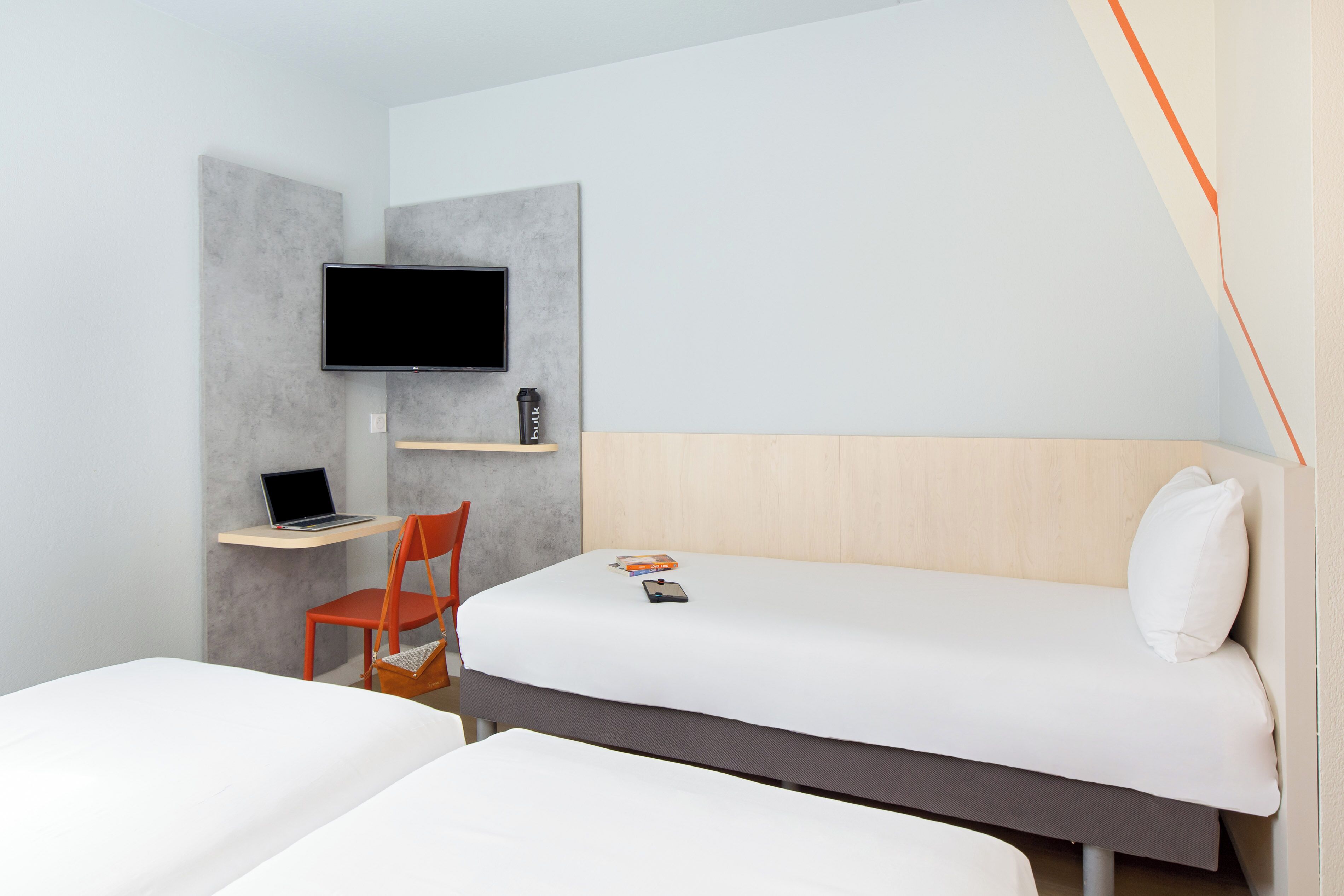 Ein Hotelzimmer mit einem Bett, einem Schreibtisch mit Laptop, einem Sessel, einem an der Wand montierten Fernseher und einem Nachttisch mit Fernbedienung.