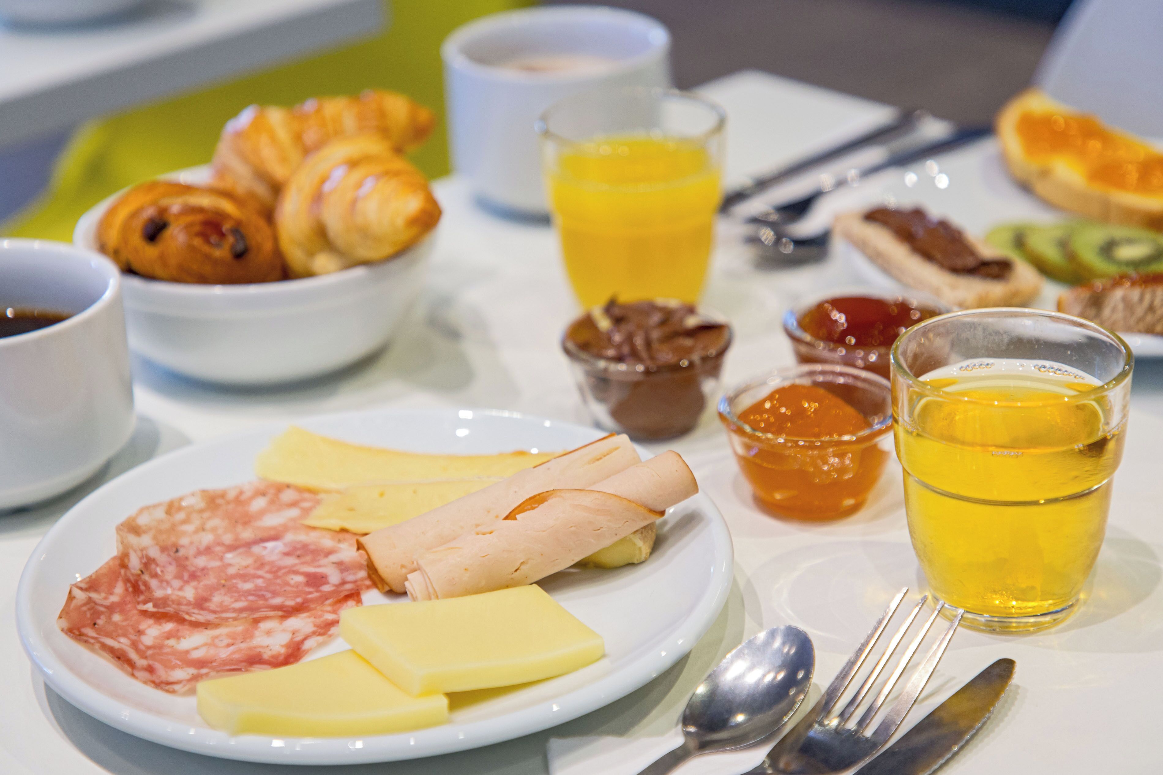 Un desayuno con croissants, queso, jamón y diversas salsas en platos, junto con vasos de zumo de naranja y un bol de pastelería.