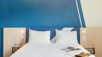 Habitación de hotel con una cama, dos almohadas blancas, una revista, una cadena dorada y una bolsa de malla negra.
