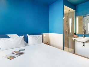 Une chambre d’hôtel avec un lit, une table de chevet, un miroir, un espace douche et un lavabo blanc.