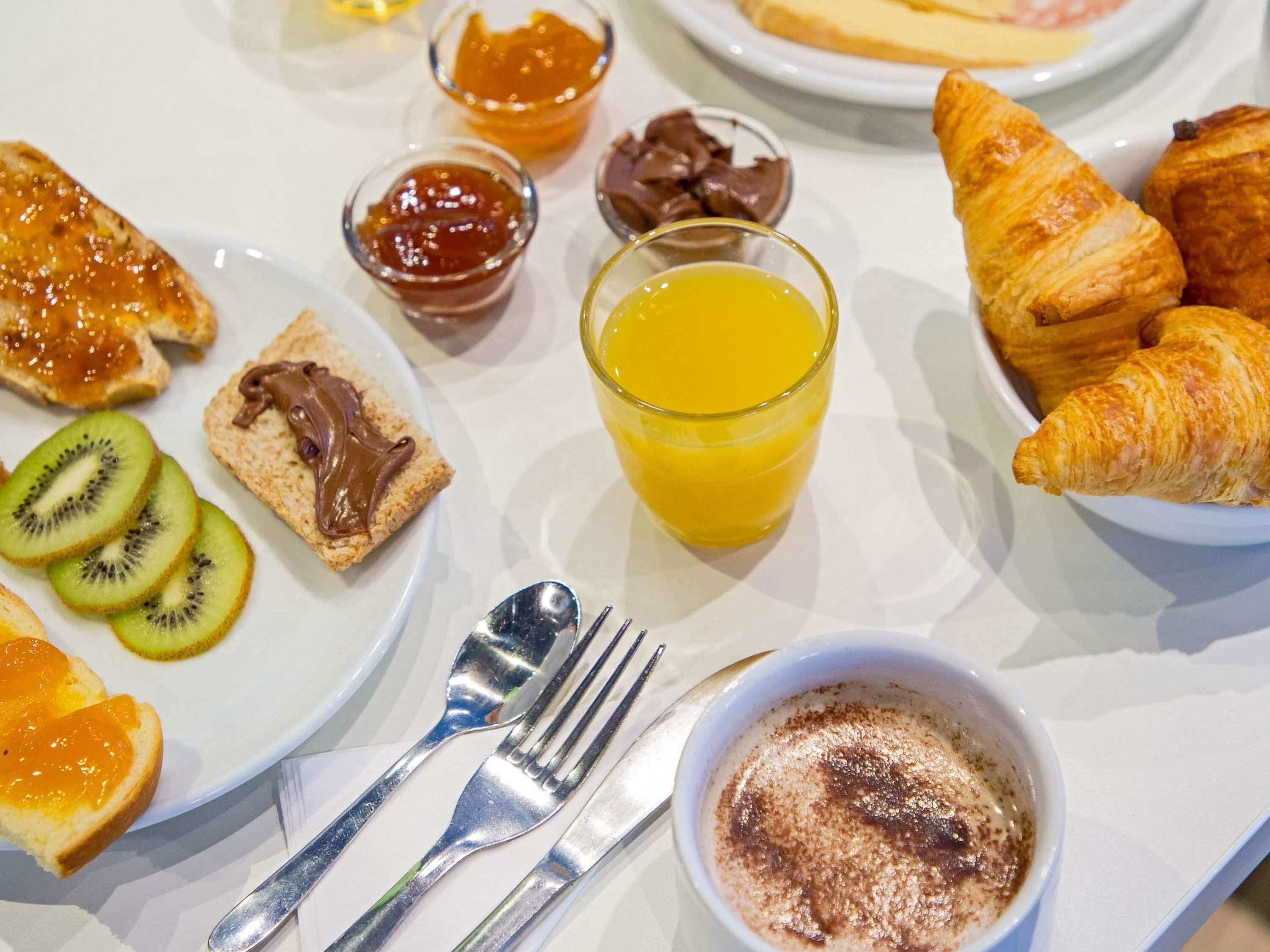 A breakfast setting with croissants, toast with spreads, kiwi slices, a glass of orange juice, and a bowl of oatmeal.