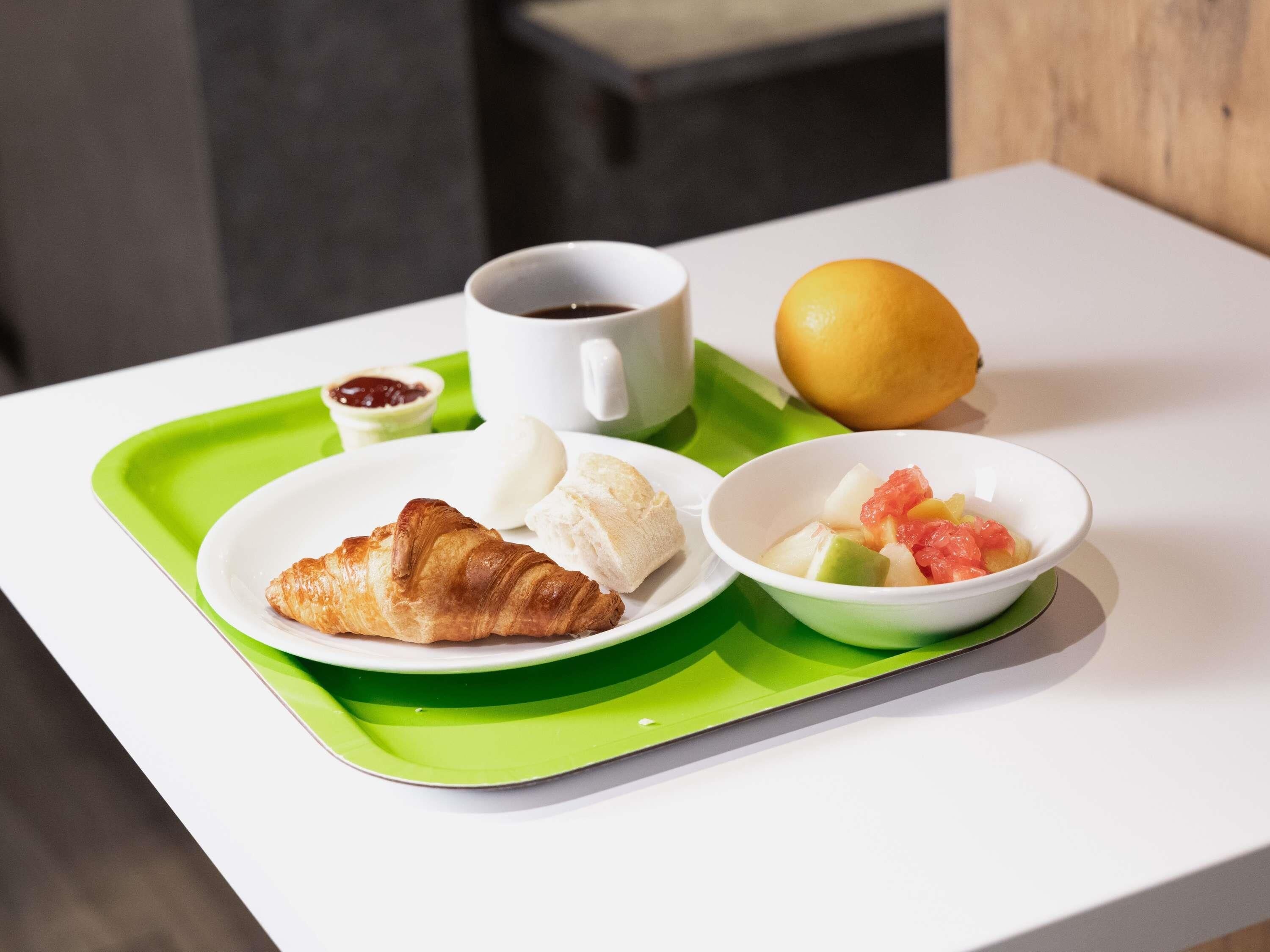 Una bandeja con un croissant, una taza de café, un limón y un cuenco con fruta.