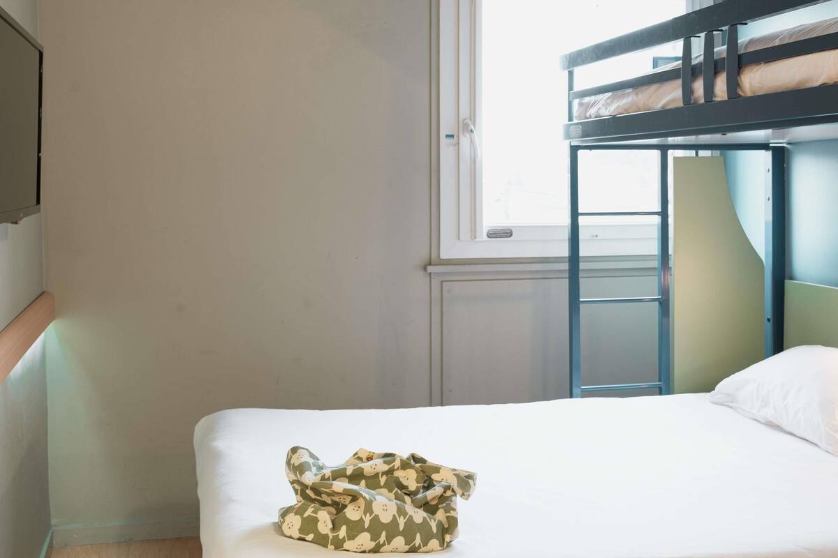 A bunk bed room with a window, a bed with a camouflage bag, and a white pillow.