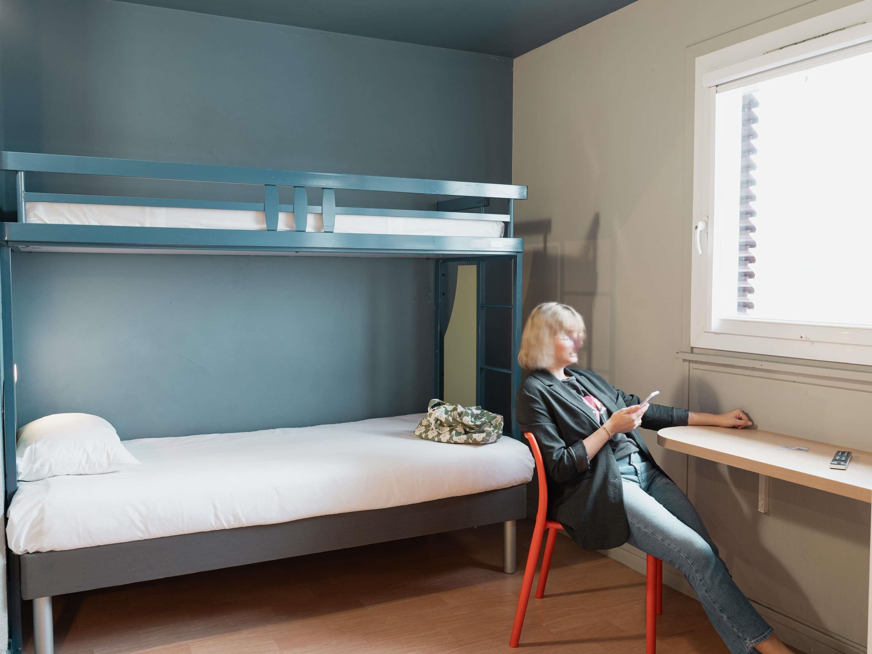 Un lit superposé avec un matelas blanc, une petite table avec une télécommande et une fenêtre avec des stores.