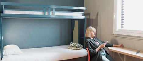Una cama litera con colchón blanco, una mesita con un control remoto y una ventana con persianas.