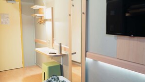 A hotel room with a bed, a green stool, a sink with a mirror, a television, and a door. - ibis budget Thonon Les Bains (Thonon-les-Bains)