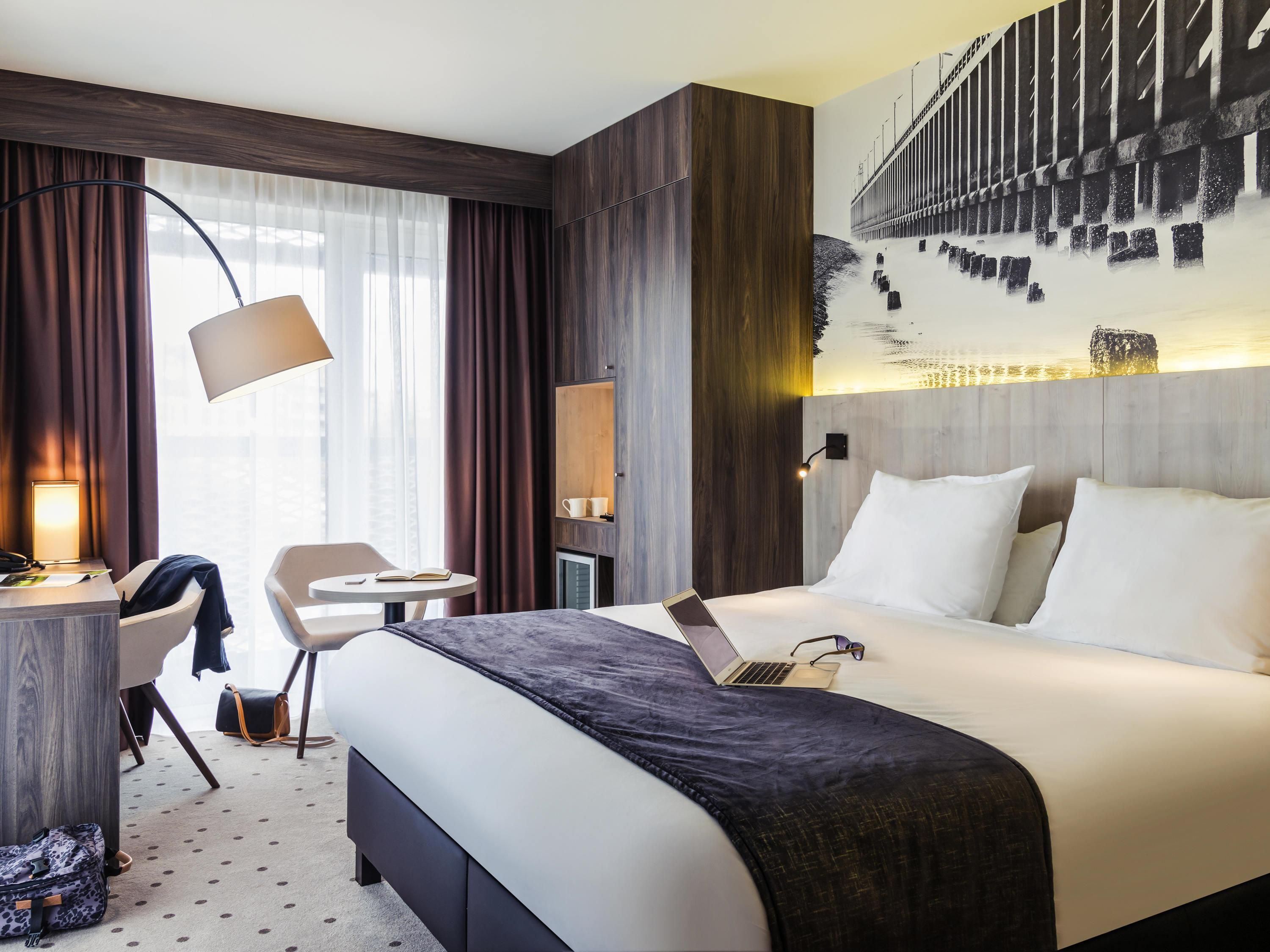 Habitación de hotel con una cama grande, un escritorio con lámpara, una silla, una computadora portátil y vista a un puente por la ventana.