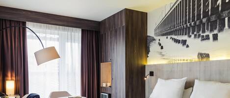 Habitación de hotel con una cama grande, un escritorio con lámpara, una silla, una computadora portátil y vista a un puente por la ventana.