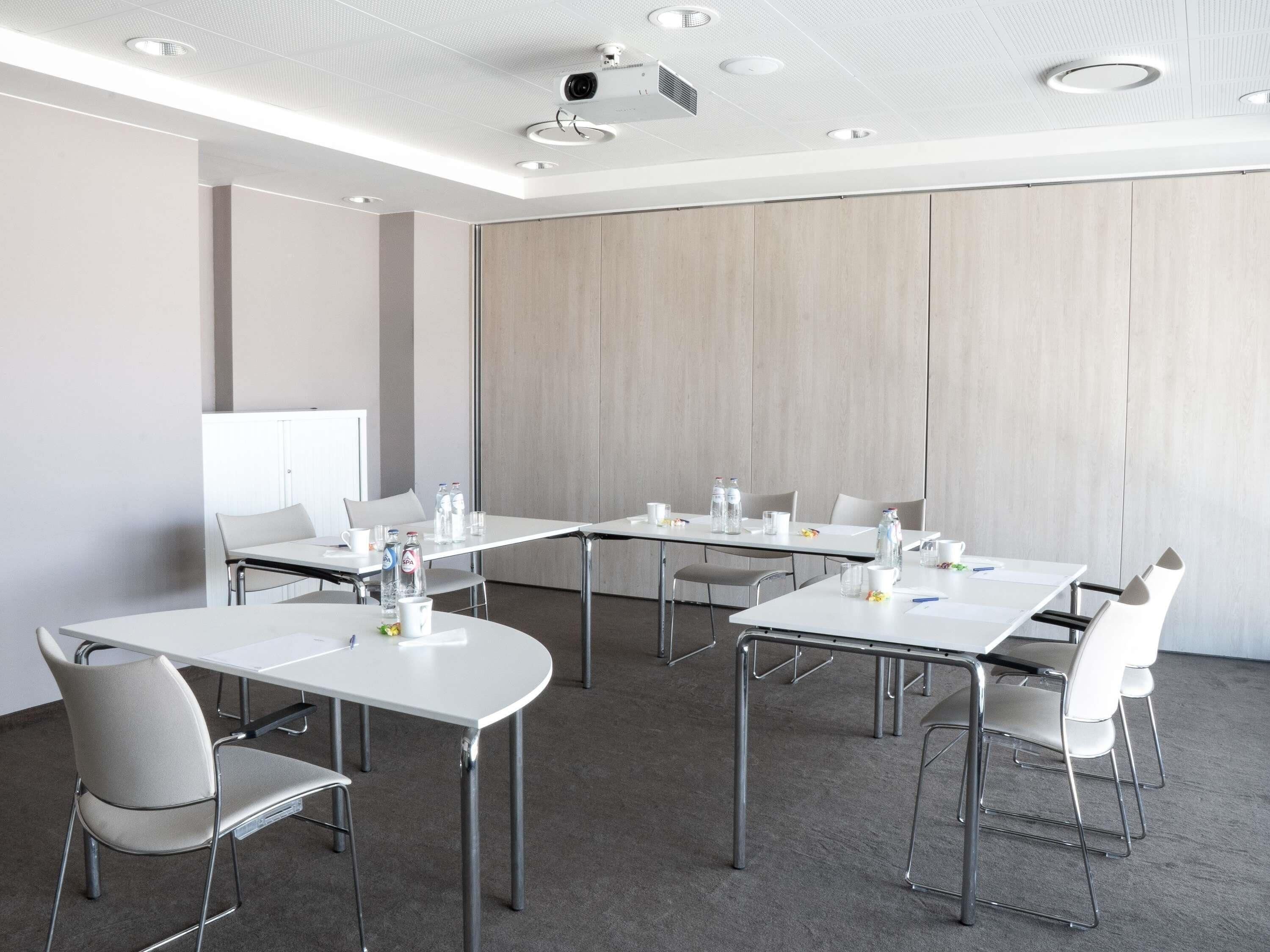 A conference room with white tables, grey chairs, a whiteboard, and a projector on the ceiling.