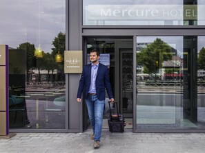 Un hombre con traje camina hacia la entrada de un hotel, donde se ve un cartel que indica 'Mercure Hotel' y 'Blankenberge'.