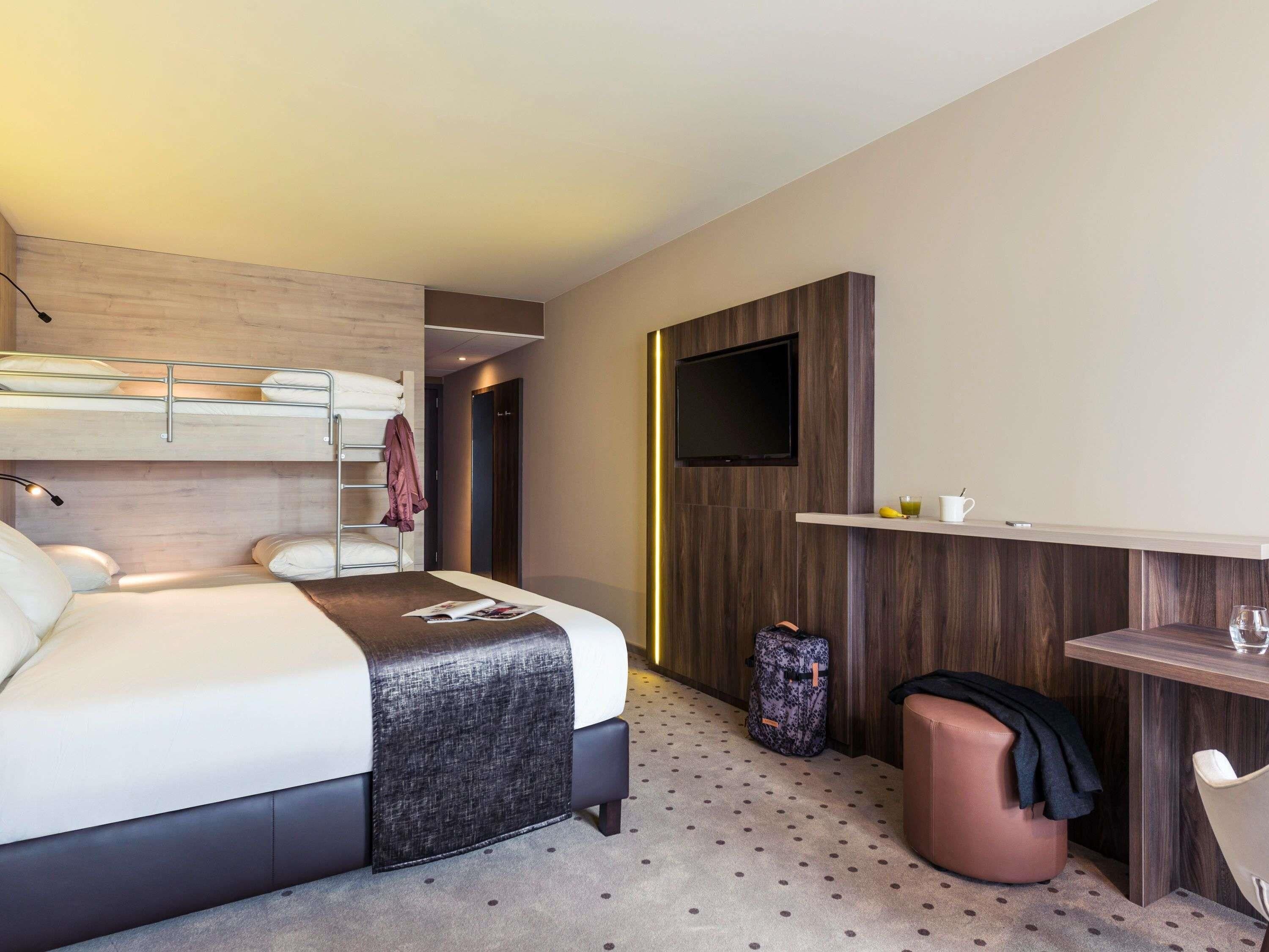 Une chambre d’hôtel moderne équipée d’un lit superposé, d’un bureau avec un téléviseur à écran plat, d’un coin salon et d’une petite kitchenette.
