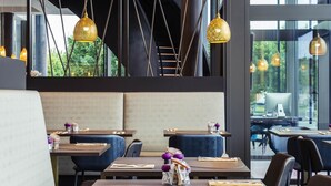 A modern restaurant interior with high tables, cushioned seating, hanging pendant lights, and a view of greenery outside.