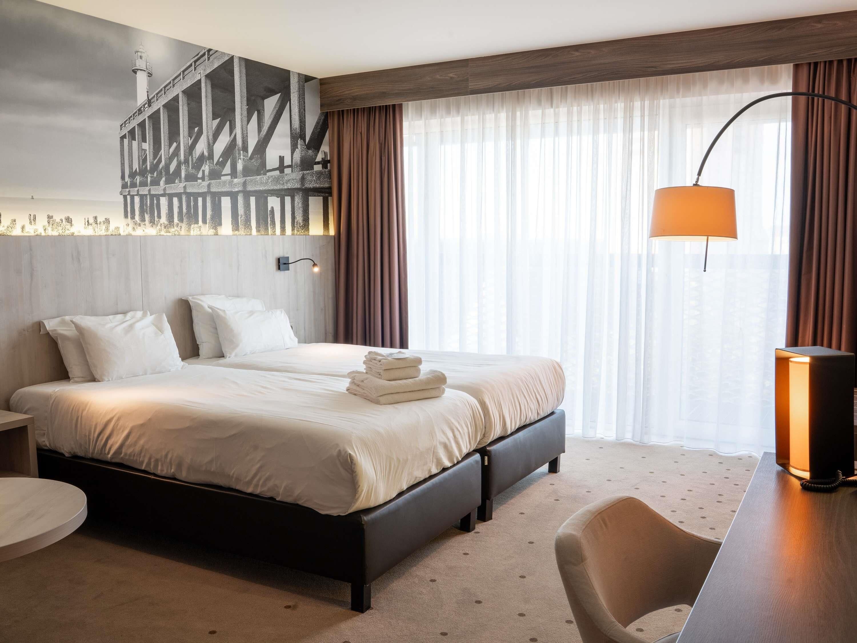 Una habitación de hotel con una cama, un escritorio, una silla, una lámpara y un ventanal con cortinas.