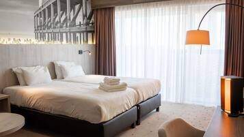 A hotel room with a bed, a desk, a chair, a lamp, and a large window with curtains.