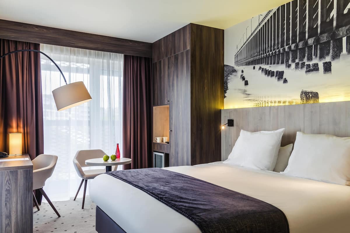 A hotel room with a bed, a desk, a chair, a small table with a bottle and apples, and a large window with curtains.