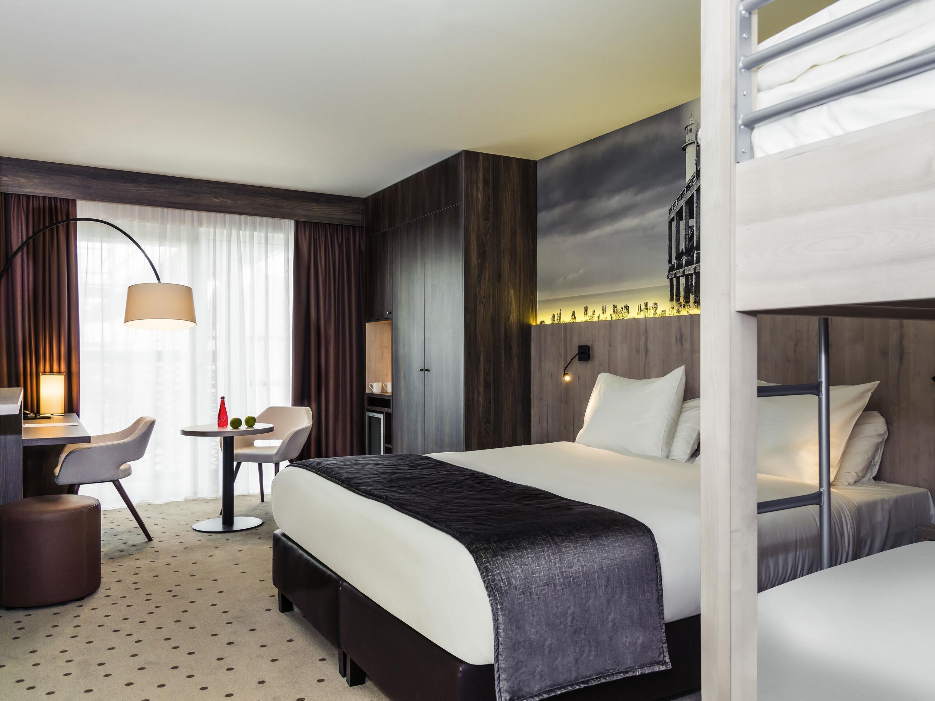 A hotel room with a bed, a desk with a chair, a small table, and a lamp.
