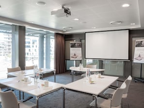 Une salle de conférence moderne, dotée de tables et de chaises blanches, d’un grand écran et d’un projecteur.
