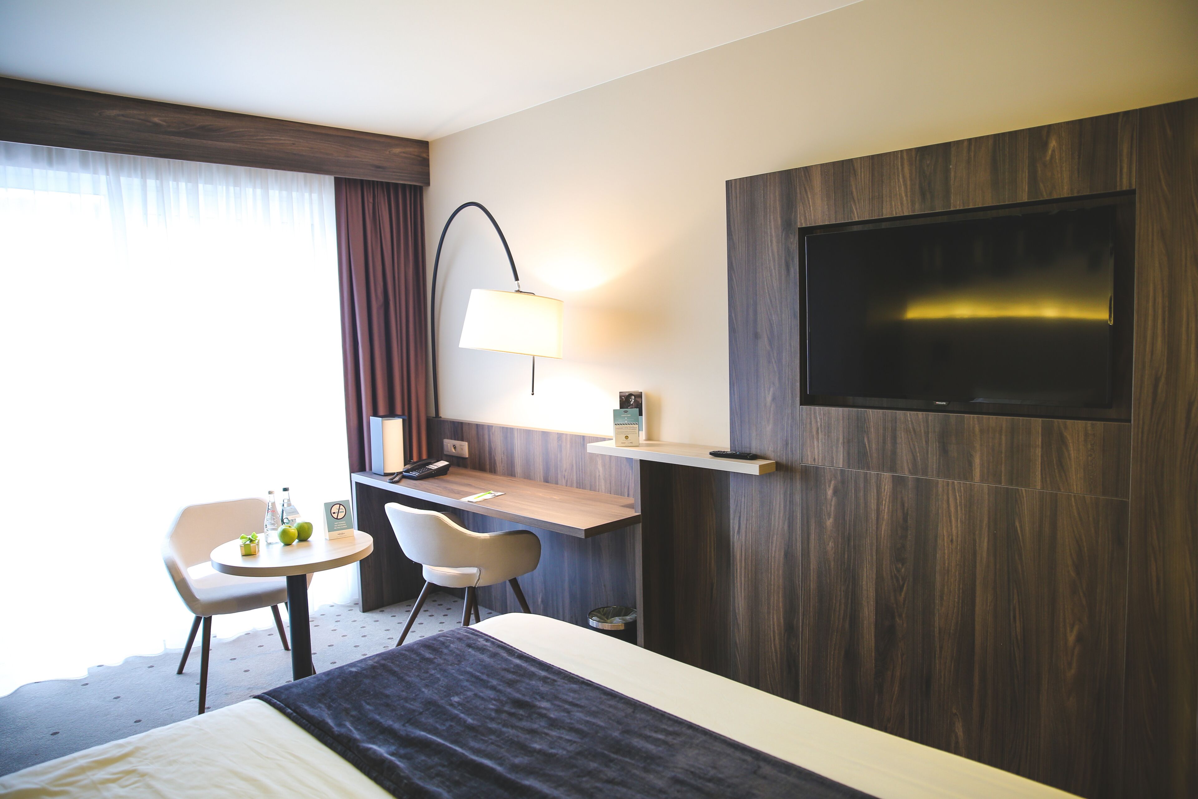 Habitación de hotel con cama, escritorio, silla, televisor en la pared y lámpara.