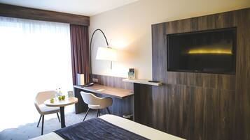 Ett hotellrum med en säng, ett skrivbord med en stol, en tv som är monterad på väggen och en lampa.
