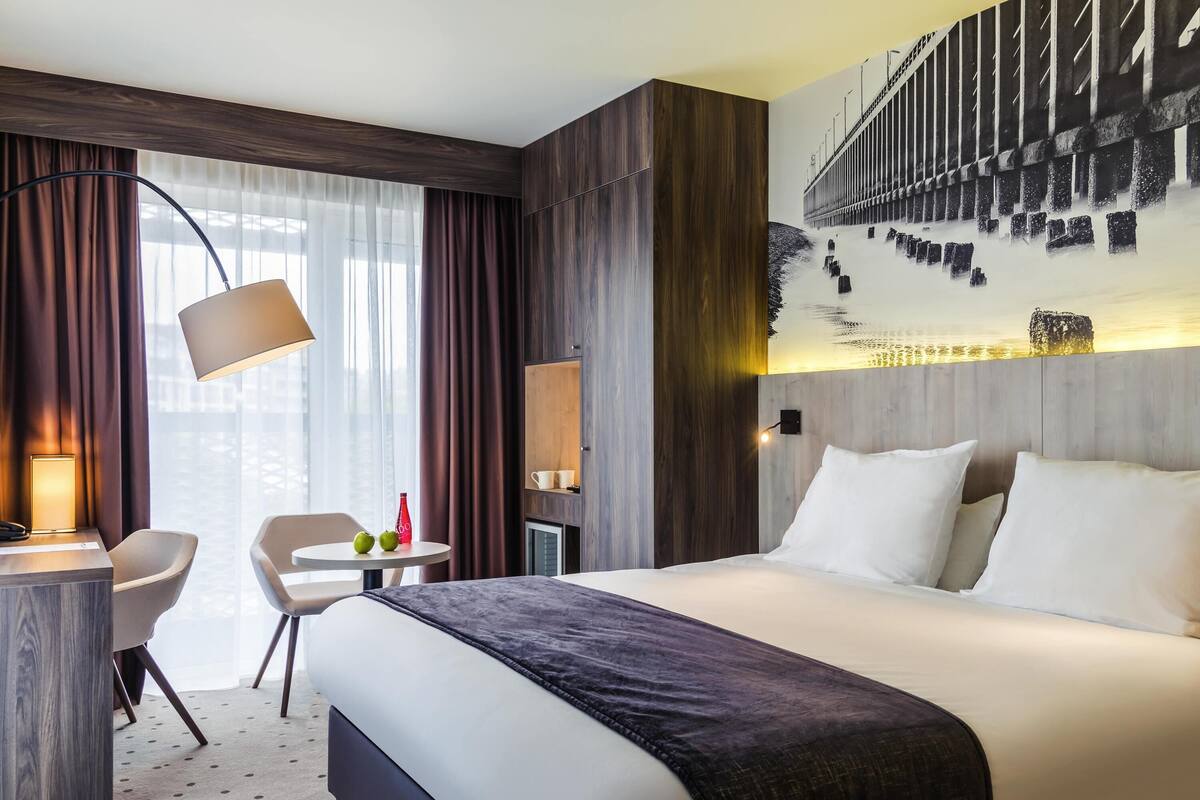Habitación de hotel con cama, escritorio, silla, una mesita con una botella y manzanas, y un ventanal con cortinas.