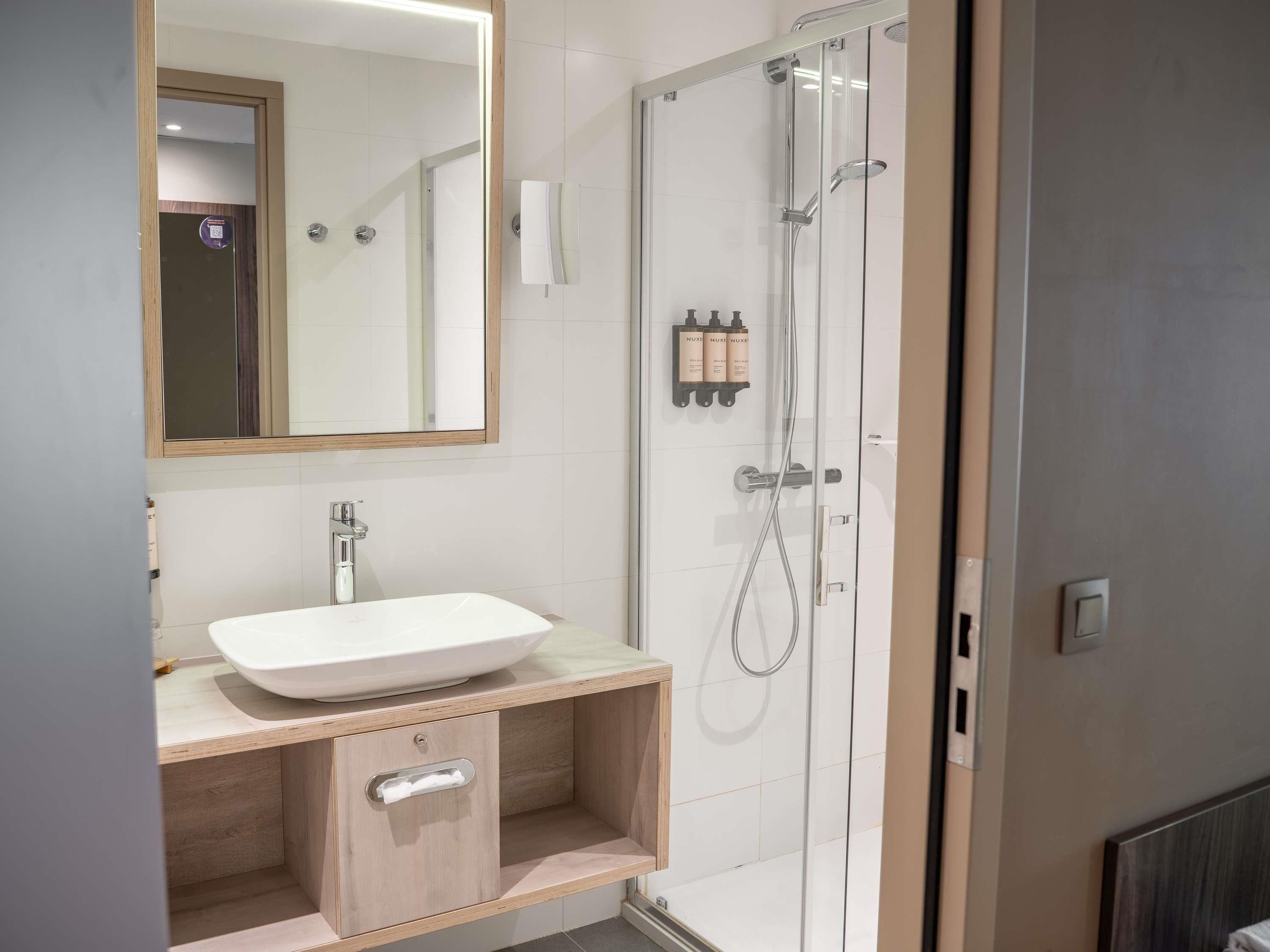 Un baño moderno con ducha envuelta en vidrio, un lavabo blanco y un mueble de tocador de madera.