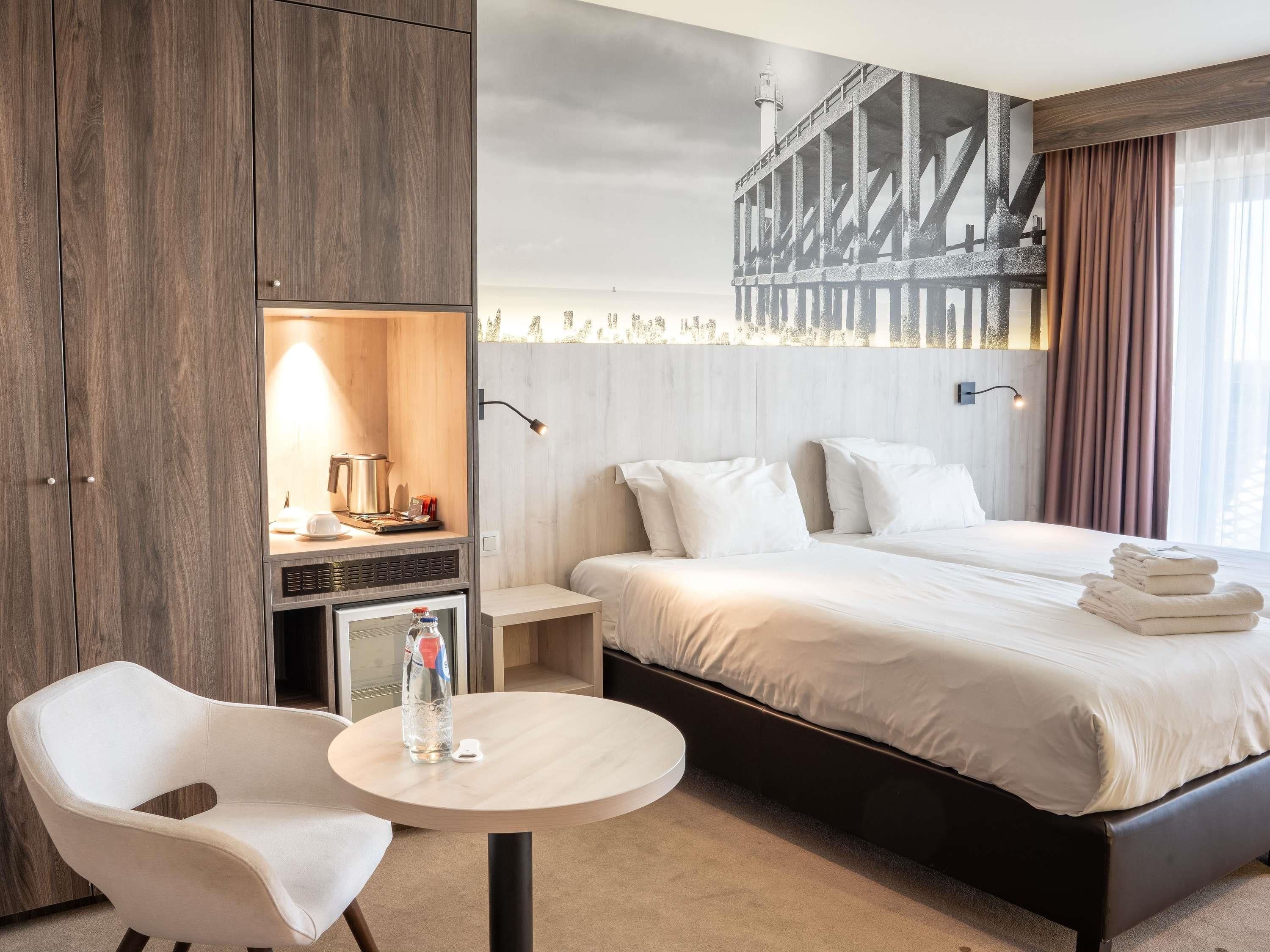 A hotel room with a bed, a desk with a lamp, a chair, and a view of a pier.