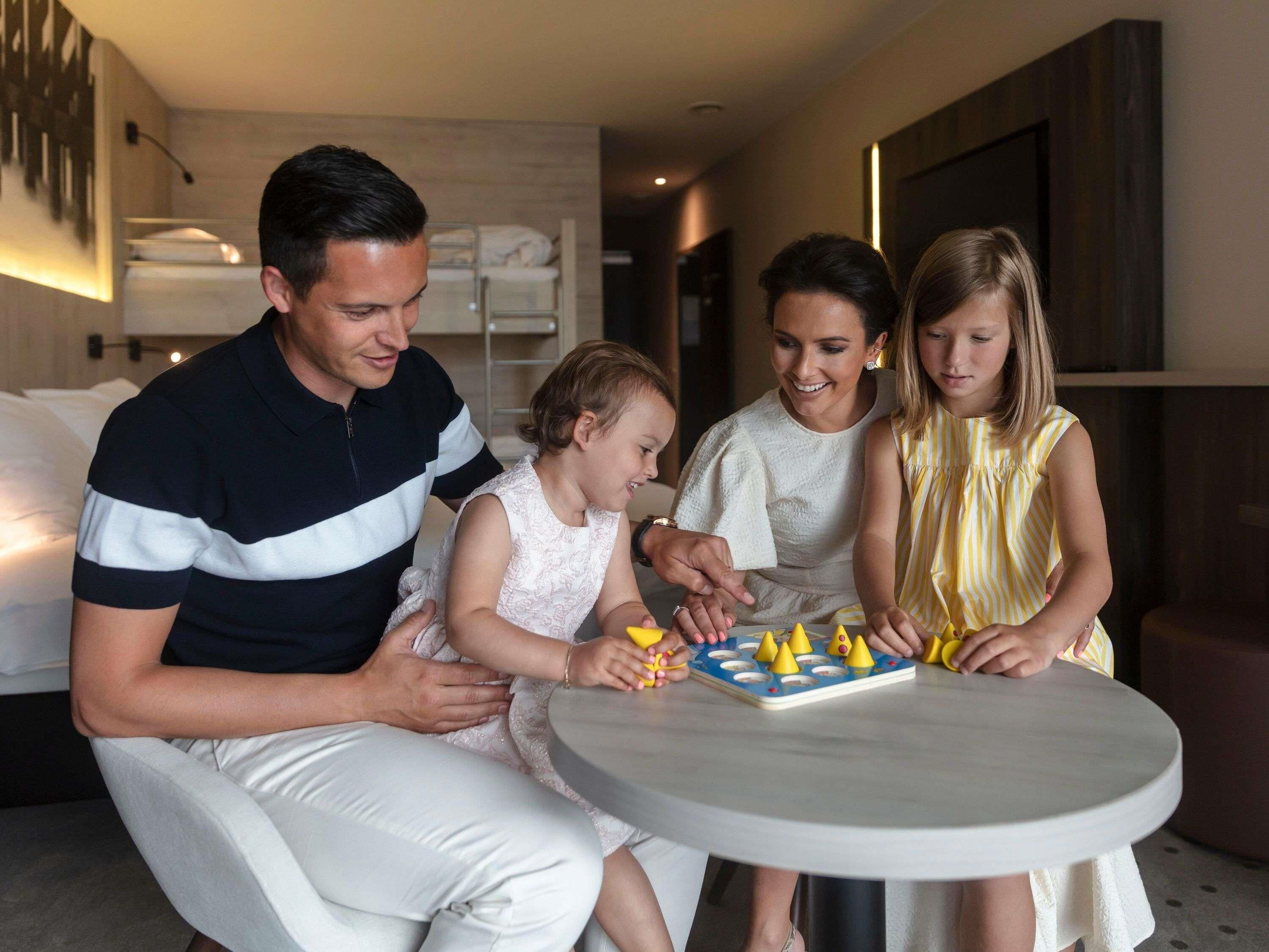 A family of four seated around a small round table with a board game, in a modern hotel room with a bed and a wall-mounted shelf in the background.