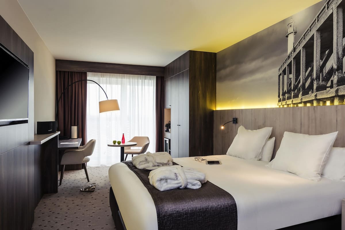 Habitació d'hotel amb llit, escriptori, cadira i una finestra gran amb cortines.